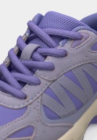 Μωβ και λιλά αθλητικό παπούτσι με ύφασμα διχτυού, επενδύσεις από σουέντ, υφασμένους πλευρικούς ρίγες και ταιριαστά μωβ κορδόνια δεμένα πάνω σε μαξιλαρωμένη γλώσσα.