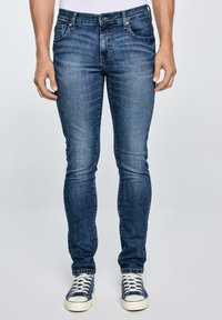 Mann in eng geschnittenen blauen Jeans und marineblauen Sneakers mit weißen Schnürsenkeln, stehend vor einem einfarbigen hellgrauen Hintergrund.