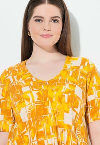 Blouse jaune à motif avec un col en V et des manches courtes, ornée de formes abstraites blanches créant un design audacieux sur un tissu texturé.