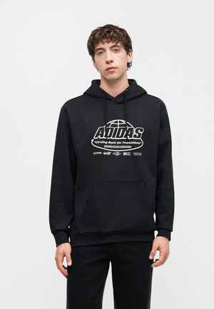 Jeune homme portant un sweat à capuche noir Adidas avec texte graphique blanc et symboles, debout devant un fond gris clair uni.