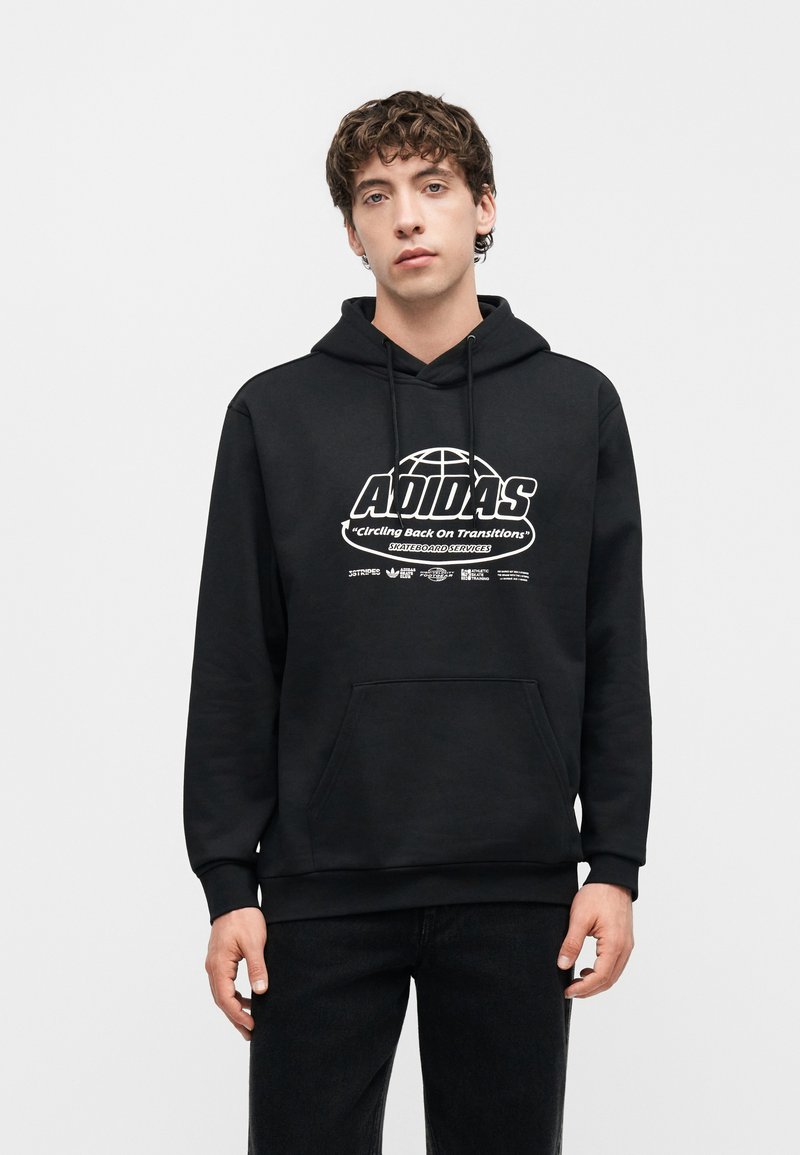Junger Mann trägt schwarzen Adidas-Hoodie mit weißem grafischem Text und Symbolen, steht vor einfachem hellgrauem Hintergrund.
