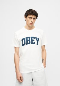 Νεαρός άνδρας με κατσαρά μαλλιά, φοράει ένα λευκό T-shirt με τη λέξη "OBEY" σε σκούρα μπλε γράμματα και ανοιχτό γκρι σορτς, μπροστά σε απλό φόντο.