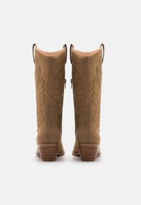 Bottes de cowboy en daim marron avec des détails en relief, un bout pointu et un talon en bois, dotées de languettes en haut pour un enfilage facile.