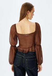 Braun transparente Bluse mit langen, gepufften Ärmeln und einem quadratischen Ausschnitt. Rüschenverzierung am Saum ergänzt das taillierte Oberteil.