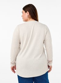Maglietta lunga a maniche con orlo arrotondato di un beige chiaro, decorata con accenti di pizzo sui polsini, realizzata in un tessuto morbido e testurizzato.
