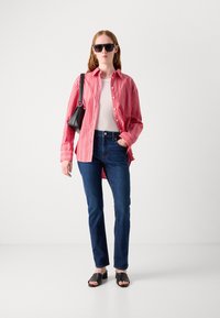 Camiseta vermelha às riscas, topo branco, jeans azul-escuro, sandálias pretas; mala preta, óculos de sol grandes e pretos; tecido suave, corte relaxado.