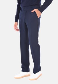 Uomo che indossa pantaloni eleganti blu navy, un maglione blu navy e sneaker bianche, in piedi con una mano in tasca contro uno sfondo bianco.