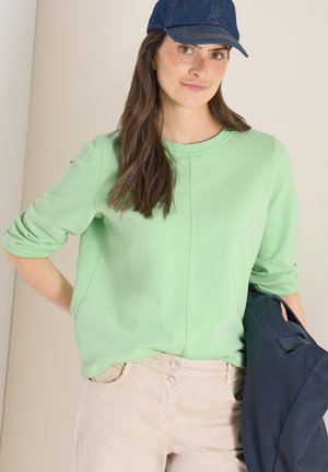 Donna con lunghi capelli scuri che indossa un berretto blu, una felpa verde chiaro e pantaloni beige, tenendo una giacca scura su un braccio.