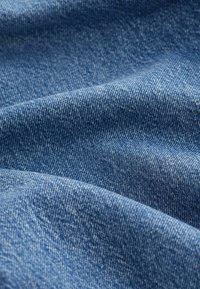 Denim fabric in medium blue, featuring a soft texture with a slight sheen. The surface shows a consistent weave pattern and subtle variations.