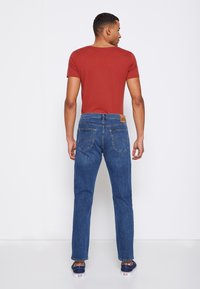 Rød t-skjorte med korte ermer kombinert med blå denimjeans. Jeansene har baklommer og et skinnlogo-patch på livet. Uformelt fottøy er synlig.
