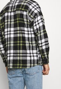 Camicia di flanella a quadri nera, bianca e verde con polsini a bottone, caratterizzata da una vestibilità rilassata e un orlo leggermente curvo. Indossata sopra jeans blu.