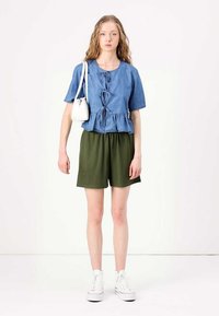 Denim peplum majica s prednjim vezanjem, uparena s olivno zelenim rastezljivim šortsem i belim tenisicama. Model nosi malu belu torbicu.