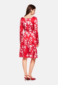 Rotes Satin-Kleid mit weißem Blumenmuster, langen Ärmeln, runder Ausschnitt und ausgestelltem Rock. Kombiniert mit roten High Heels.