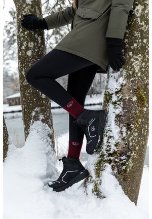 Black snow boots with reflective accents, paired with burgundy socks featuring a logo, worn with black leggings and a green jacket in snow.