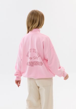 Osoba o jasnobrązowych włosach w różowej bluzie z tekstem na plecach i kremowych spodniach, stojąca na jednolitym białym tle.