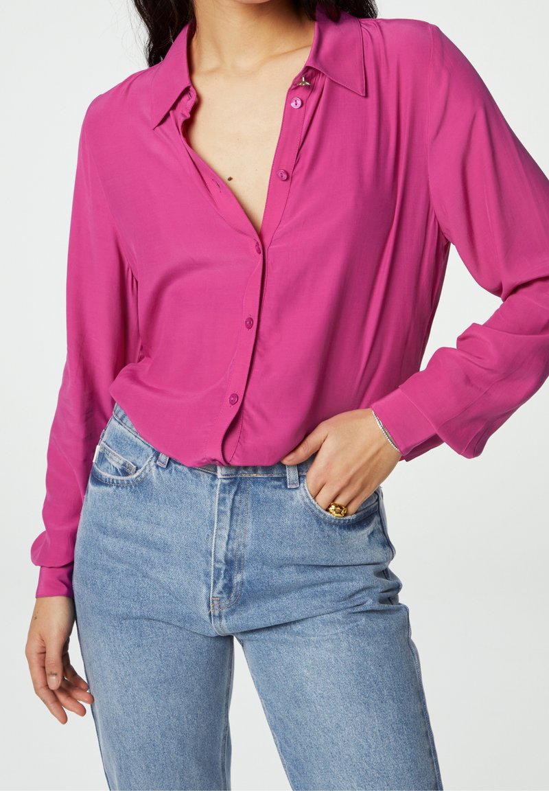 Femme portant un chemisier rose vif boutonné rentré dans un jean bleu clair taille haute, la main posée dans la poche et une bague en or visible.