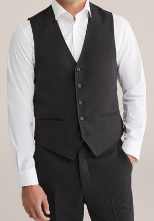 Mann trägt schwarze, geknöpfte Weste über weißem Hemd mit schwarzen formellen Hosen, Hand in der Tasche, neutraler Hintergrund.