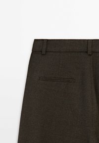 Pantalon marron foncé sur mesure montrant la taille avec passants de ceinture et une seule poche arrière horizontale passepoilée sur fond blanc.