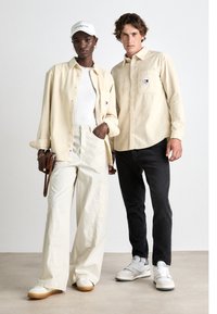 Camisa de pana beige, top blanco de canalé, pantalones anchos crema, vaqueros negros y zapatillas blancas. Los accesorios incluyen una gorra blanca y un bolso marrón.
