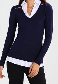 Frau trägt einen marineblauen V-Ausschnitt-Pullover mit Schulterreißverschlüssen über einem weißen Hemd mit Kragen und schwarzen Hosen, steht vor einem weißen Hintergrund.