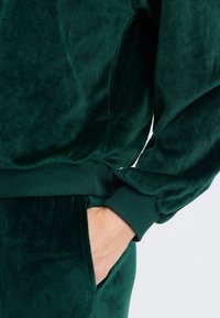 Felpa in velluto verde con polsini e vita a costine, caratterizzata da una texture liscia e una mano nella tasca laterale.