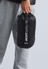 Svart packväska med dragsko i toppen, märkt "JACKIT SATEL1TE" i vit text, hålls i en hand och visar sitt släta, lätta material.
