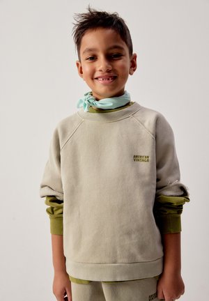Niño sonriente que lleva una sudadera beige de "American Vintage" sobre una camisa verde, con una bufanda azul claro atada alrededor del cuello.