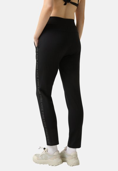 Zwarte joggingbroek met een hoge taille, zijzakken en een verticale reflecterende logo langs het buitenbeen, gecombineerd met lichtgekleurde sneakers.