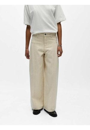 Person iført beige vide bukser med vertikale sømmer, en hvit kortermet skjorte, svarte sko og et sølvarmbånd, stående mot en hvit bakgrunn.