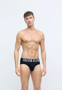 Čierne elastické nohavičky so širokým bielym pásom s logom "CALVIN KLEIN", hladká textura, navrhnuté pre tesný strih.