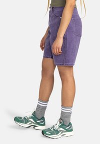 Shorts di denim viola con una vestibilità dritta, passanti per la cintura e tasche frontali, abbinati a scarpe da ginnastica verdi e calzini a strisce grigie.