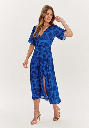 Kobieta w niebieskiej kwiecistej sukience midi z dekoltem w serek i bocznym rozcięciem, pozująca z jedną ręką blisko głowy i złotymi sandałami na wysokim obcasie.