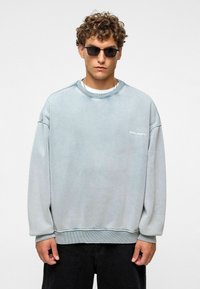 Lichtblauwe oversized sweatshirt van zachte stof, met een geribbelde halslijn en manchetten, voorzien van een wit logo op de borst. Draag het met een zonnebril.