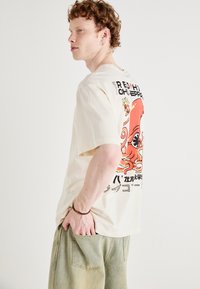 Beige oversized t-shirt med et farverigt blæksprutteprint på ryggen, med sort tekst og en afslappet pasform. Let bomuldsstof.