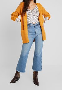 Kvinna i orange stickad kavaj, rutigt blus, korta blåjeans och bruna spetstövlar mot en enfärgad bakgrund.