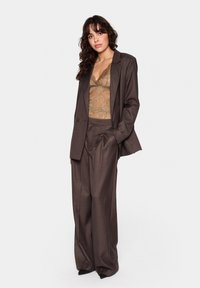 Un costume marron sur mesure avec une coupe ample, des jambes larges et un blazer à un bouton. En dessous, un top en dentelle marron avec un décolleté en V.