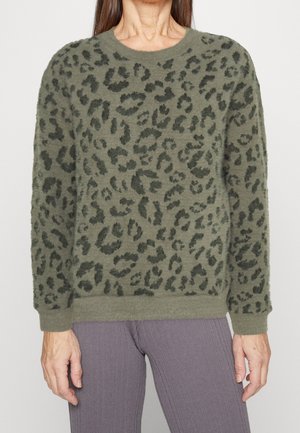 Femme portant un pull duveteux vert olive avec des taches léopard foncées et des poignets côtelés, associé à un pantalon violet côtelé.