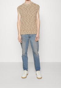 Tant stickad ärmlös topp med ett mönster, kombinerad med ljusblå denimjeans och vita sneakers med tan-färgade accenter.