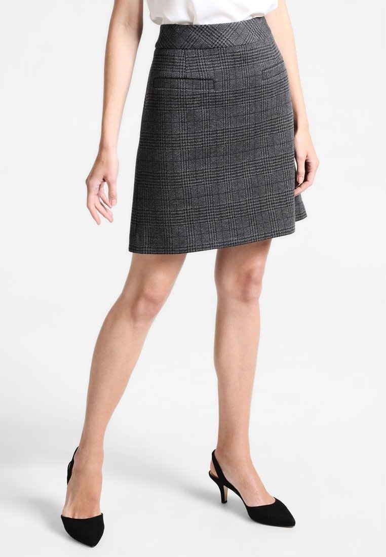 Mini jupe en tartan gris en tissu texturé, dotée de deux poches avant et d'un ourlet évasé. Portée avec des chaussures à talons noirs.