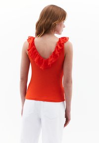 Top rosso senza maniche con scollatura in pizzo arricciato, texture a costine e forma aderente. Abbinato a pantaloni bianchi a vita alta.