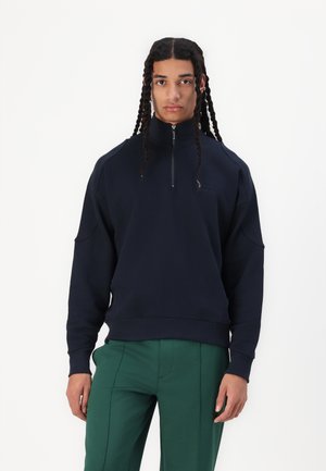 Navy blue, zip-up sweatshirt with high collar, dropped shoulders, and ribbed cuffs. Fabric has a smooth texture; subtle logo detail.