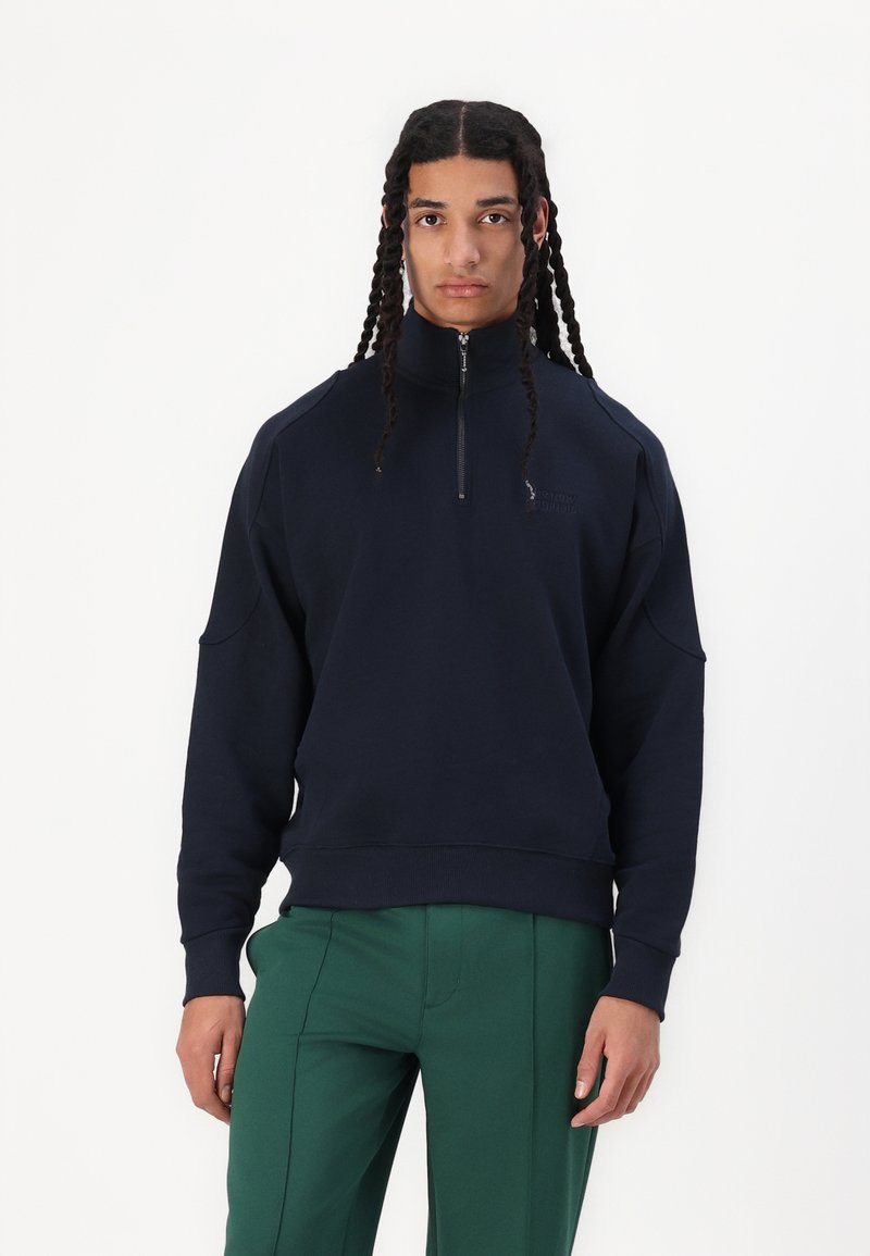 Marineblå, glidelås-genser med høy krage, senkede skuldre og ribbestrikkede mansjetter. Stoffet har en glatt tekstur; subtil logo-detajl.