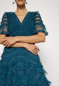 Vrouw draagt een donkergroene jurk met ruches, doorschijnende gestreepte korte mouwen en hangende oorbellen, armen gekruist ter hoogte van de taille.