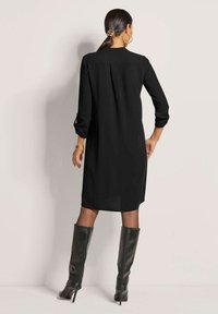 Schwarzes langärmeliges Kleid mit lockerer Passform, welches einen Rückenplissee und hochgerollte Ärmel hat, kombiniert mit hohen schwarzen Stiefeln mit Absatz.