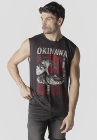 Hombre con camisa negra sin mangas con el texto "OKINAWA" y un gráfico de ola, mirando hacia un lado, de pie contra un fondo claro y liso.