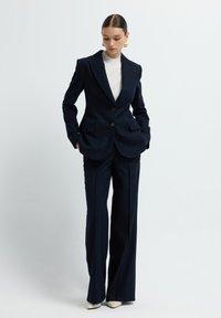 Abito blu navy con righe sottili, composto da un blazer su misura con due bottoni, pantaloni a gamba larga e una maglietta bianca aderente sotto. Scarpe con tacco completano il look.