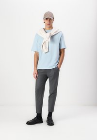 Světle modrá košile s krátkým rukávem v kombinaci se šedými tapered kalhotami, krémový svetr uvázaný na ramenou a černé slip-on boty.