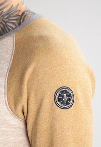 Långärmad skjorta i beige och senapsgul färgskala, med en grå krage och en cirkulär svart tygpatch på överarmen.