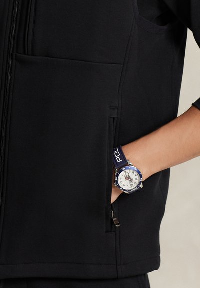 Μαύρο μπουφάν με φερμουάρ και τσέπη στην αριστερή πλευρά. Μπλε και λευκό ρολόι με ασημένιο στεφάνι και λαστιχένιο λουράκι που ξεπροβάλλει από την τσέπη.