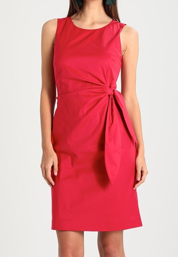 Robe rouge sans manches longueur genou avec un col rond et un détail en tissu noué à la taille sur une personne debout.